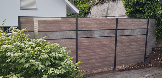 Sichtschutzzaun in brauner Holzoptik mit grauem Dekorband und dunklen Pfosten neben einem Wohnhaus.