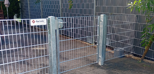 Gartentor aus Metall mit Doppelstabmattenzaun vor einem grauen Sichtschutzzaun am gepflasterten Garteneingang.