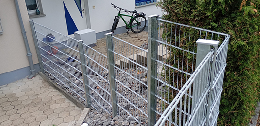 Metall-Gitterzaun mit rechteckigen Maschen und silberfarbenen Pfosten entlang eines Kiesbeets am Hauseingang.