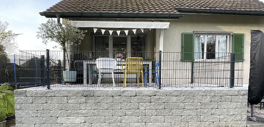Anthrazitfarbener Doppelstabmattenzaun auf einer Steinmauer zur Einfassung einer erhöhten Terrasse am Wohnhaus.