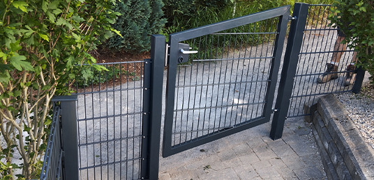 Anthrazitfarbenes Gartentor mit Gitterfüllung und passendem Doppelstabmattenzaun zwischen bepflanzten Gartenbereichen.