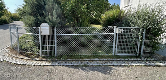 Maschendrahtzaun mit Gartentor und Briefkasten entlang eines begrünten Grundstücks an einer Strasse.